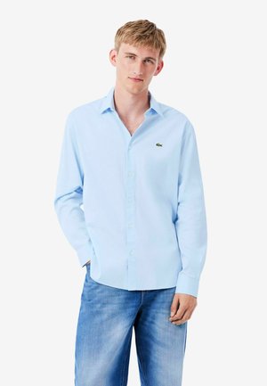Joven con una camisa azul claro abotonada con un pequeño logo verde y vaqueros azules de lavado medio, de pie con una mano en el bolsillo.