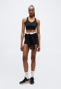 Femme athlétique portant un soutien-gorge de sport noir, un short, une veste noire nouée à la taille, des chaussettes blanches Nike et des chaussures de course noires, debout face à l'avant.