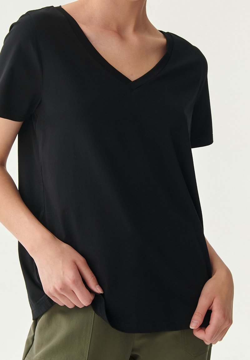 Personne portant un T-shirt noir à manches courtes et col en V, et un pantalon vert olive, les mains reposant près des hanches.