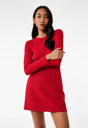 Rød ribbet langærmet top parret med en struktureret rød mini nederdel. Outfittet har tætsiddende former og et glat, blødt udseende.