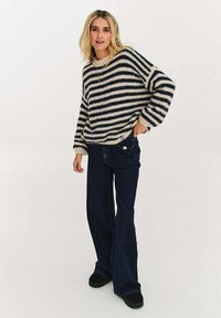 Pruhovaný svetr v krémové a námořnické modři, volného střihu s nadměrnými rukávy, doplněný tmavě modrými rozšířenými džíny a černými botami.