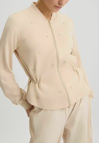 Veste beige à fermeture éclair avec col montant, arborant une taille élastique, des accents dorés décoratifs et une texture lisse.