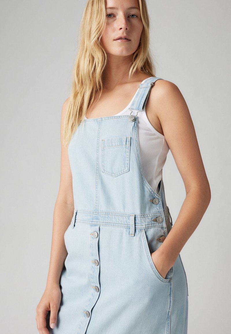 Hellblaues Jeanskleid mit verstellbaren Trägern, Vordertasche und Knopfverschluss. Glatte Textur und lässiges Design. Klassischer Overall-Stil.