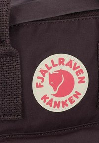 Zaino in tessuto color marrone scuro con logo circolare crema ben visibile, che presenta una volpe rosa e la scritta "FJÄLLRÄVEN KÄNKEN". Si notano il rinforzo delle cuciture e delle cinghie.