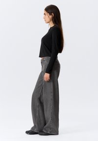 Schwarzes, langärmeliges Crop-Top kombiniert mit hoch taillierter, weit geschnittener grauer Jeans aus Denim. Der Stoff weist eine dezente Textur auf, dazu werden schwarze Schuhe getragen.