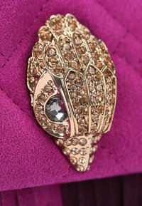 Spilla in tono oro a forma di mano, decorata con strass trasparenti e marroni chiari, posizionata su uno sfondo di tessuto viola testurizzato.