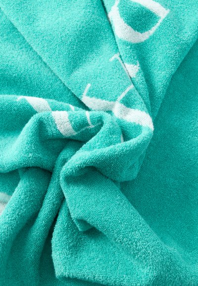 Πετσέτα από πετσετέ ύφασμα σε τυρκουάζ χρώμα με λευκά γράμματα, διπλωμένη και στριμμένη ώστε να σχηματίζει έναν απαλό, υφασματώδη κόμπο.