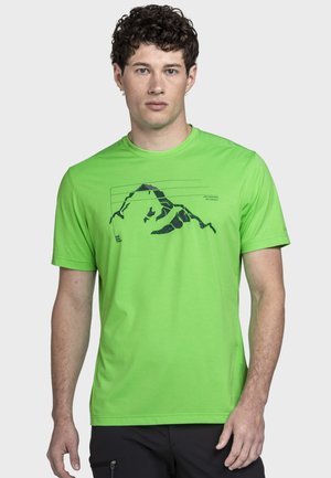 Mann trägt ein leuchtend grünes Kurzarmshirt mit schwarzer Berggrafik und Text, steht vor einem schlichten hellen Hintergrund.