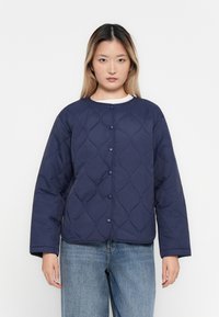 Frau in einer marineblauen gesteppte Jacke mit Druckknöpfen, die über einem weißen Oberteil und hellblauen Jeans steht, vor einem schlichten weißen Hintergrund.