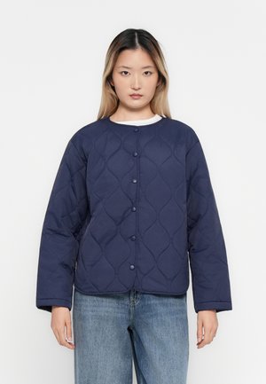 Woman wearing a navy quilted jacket with snap buttons over a white top and light blue jeans, standing against a plain white background.
