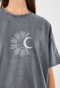 Graues Baumwoll-T-Shirt mit einem weißen Grafikdesign eines Halbmonds mit Sonnenstrahlen und dem Text "STEH AUF UND STRAHL." Lockere Passform, kurze Ärmel.