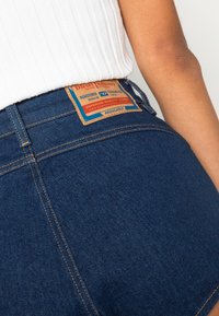 Los vaqueros de mezclilla azul oscuro presentan una cintura alta, costuras visibles y un parche de cuero con la marca en la parte posterior.