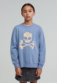 Hanorac albastru deschis cu un grafic floral de craniu și oase încrucișate. Guler și manșete ribbate, textură moale. Asortat cu o fustă din denim gri.