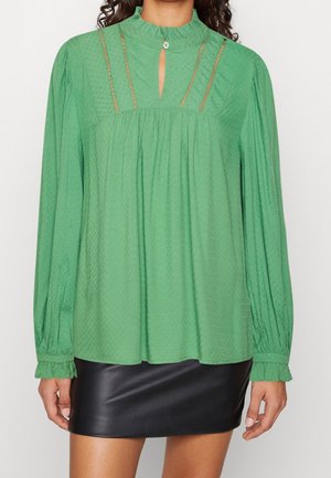 Kvinde iført en grøn langærmet bluse med blondeindre, flæsekant ved halsen og ærmekanter, kombineret med en sort lædernederdel.