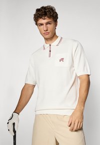 Hvit strikket polo skjorte med krage, rød og hvit stripet kant, lomme med broderte initialer og korte ermer.