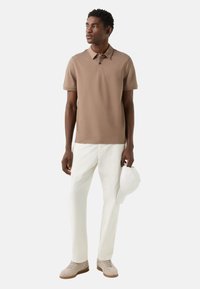 Beżowa koszulka polo, białe spodnie, beżowe trampki. Gładka tkanina, klasyczny kołnierzyk, krótkie rękawy, kapelusz trzymany w ręku, minimalistyczny styl.