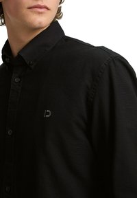 Camisa negra de botones con cuello, que presenta un logo sutil en el pecho, confeccionada con una tela texturada y un corte ajustado.
