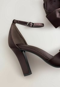 Sandale à talon haut brun foncé avec bride cheville et boucle, matière satinée, et talon bloc sur fond clair.