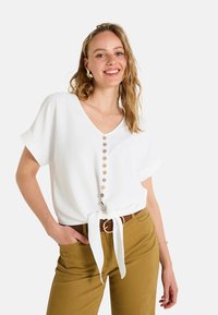 Top blanco de manga corta con escote en V, que presenta una fila de botones de madera y un detalle de cintura atada, complementado con pantalones de color caqui.