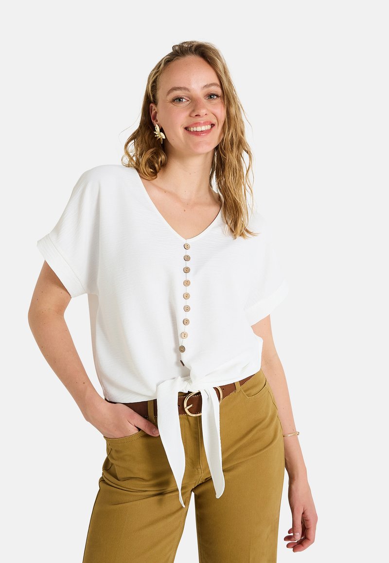 Top blanco de manga corta con escote en V, que presenta una fila de botones de madera y un detalle de cintura atada, complementado con pantalones de color caqui.