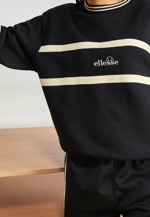 Personne portant un sweat-shirt ellesse noir avec des rayures horizontales crème et un pantalon noir assorti avec des bandes latérales vertes.