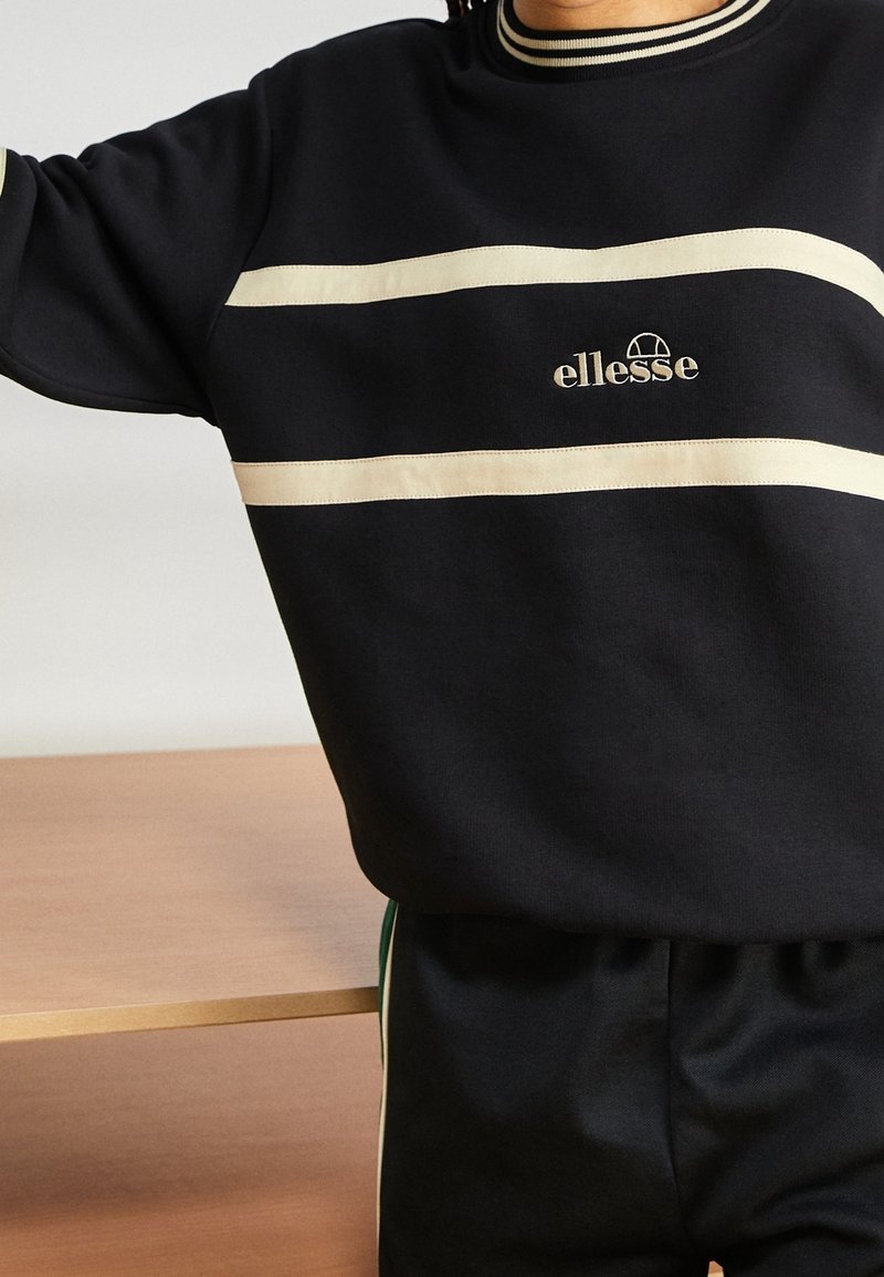 Personne portant un sweat-shirt ellesse noir avec des rayures horizontales crème et un pantalon noir assorti avec des bandes latérales vertes.
