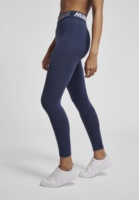 Leggings blu con vita alta e logo, realizzati in materiale elastico. Abbinati a scarpe sportive bianche. Texture liscia, design aderente.