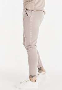 Pantalones de chándal de un beige claro con tejido texturizado, corte ajustado, bolsillos laterales y detalle del logo en el muslo. Combinados con zapatillas blancas.