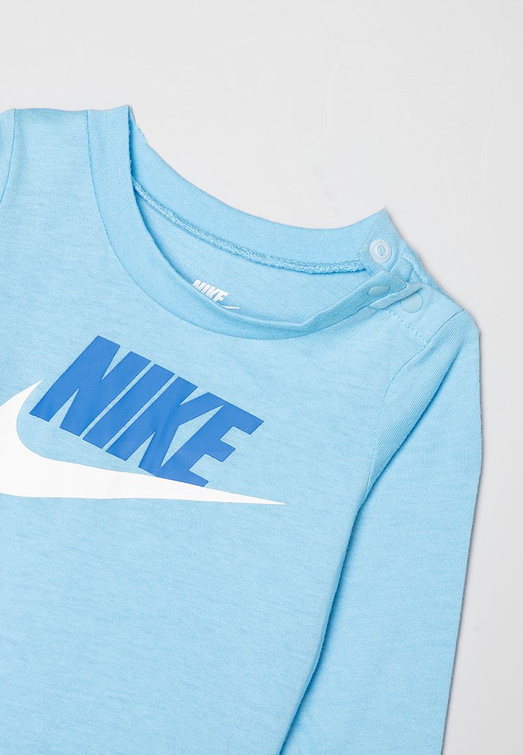 Chemise à manches longues bleu clair en tissu doux, ornée d'un grand logo "NIKE" bleu et d'un swoosh blanc. Boutons-pression près du col.
