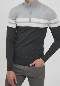 Mann in einem Langarm-Half-Zip-Pullover mit hellgrauen, weißen und dunkelgrauen horizontalen Streifen, kombiniert mit grauen Hosen.
