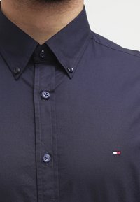 Tmavě námořnická košile na knoflíky s klasickým límcem, se světle modrými knoflíky a malým červeno-bílo-modrým logem na hrudi.