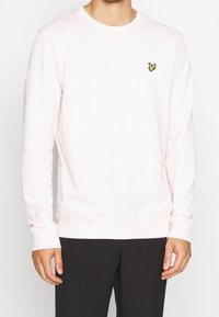 Felpa crewneck rosa chiaro realizzata in tessuto morbido, con un piccolo logo giallo ricamato sul lato sinistro del petto e polsini e orlo a costine.