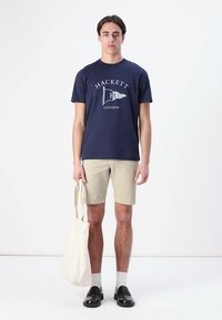 Camiseta de manga corta azul marino con un logo gráfico, combinada con pantalones cortos beige, calcetas blancas y zapatos negros sin cordones. Llevando un bolso de mano beige.