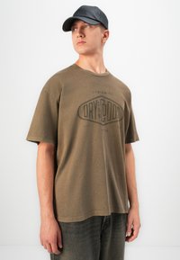 Ung mann iført en brun "Dry Goods" t-skjorte, svart caps og mørke jeans, som står mot en enkel lys bakgrunn og ser sidelengs.