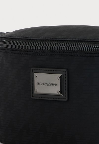 Κοντινή λήψη μαύρης τσάντας Emporio Armani με μεταλλική πλακέτα λογότυπου και εμφανές φερμουάρ κατά μήκος της πάνω άκρης.