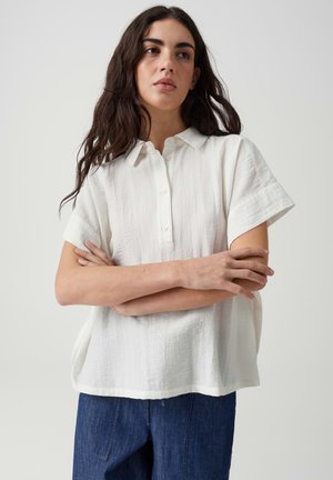 Jeune femme aux longs cheveux foncés portant une chemise blanche ample à manches courtes et un pantalon bleu, debout les bras croisés devant un fond uni.