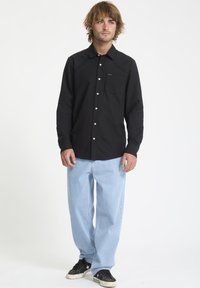 Chemise noire à manches longues avec un col à bouton, poche avant et boutons blancs, associée à un jean large bleu clair et des baskets noires.