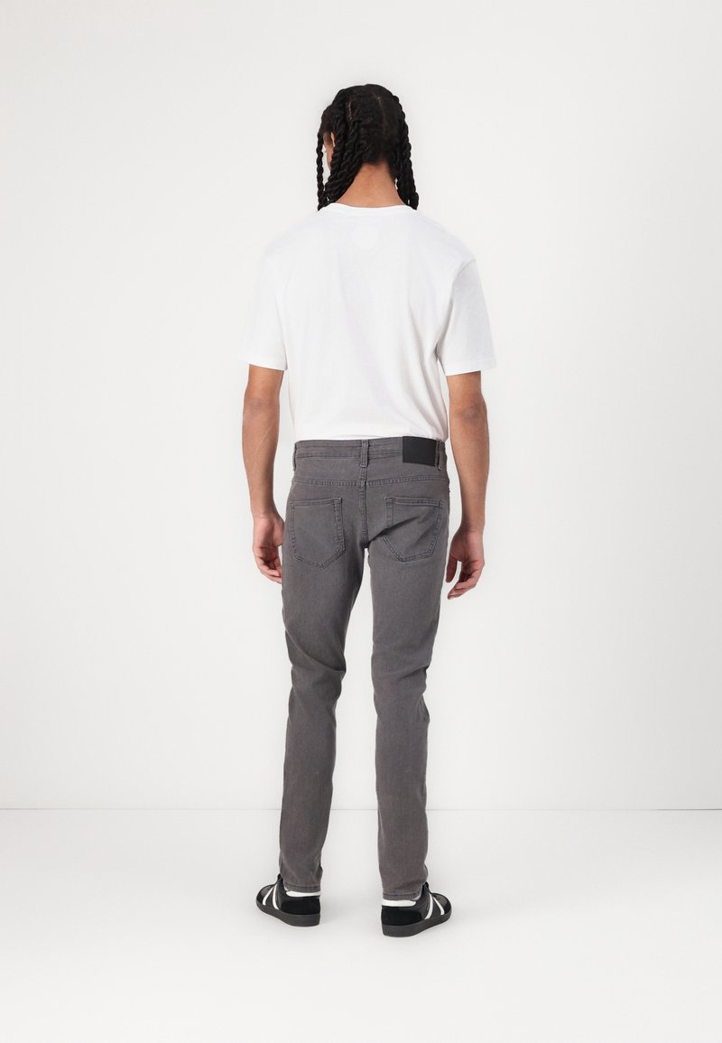 Personne avec des cheveux tressés vue de dos, portant un t-shirt blanc, un jean gris et des baskets noires dans un espace blanc et simple.
