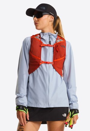 Giacca a vento azzurro chiaro abbinata a un gilet da corsa arancione con cinghie regolabili e tasche, indossata con un berretto nero e guanti.