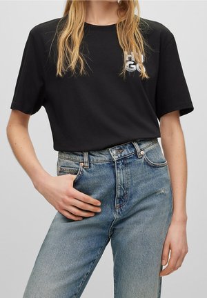 Person iført en sort Hugo t-shirt stoppet ned i højtaljede blå jeans med den ene hånd i lommen.