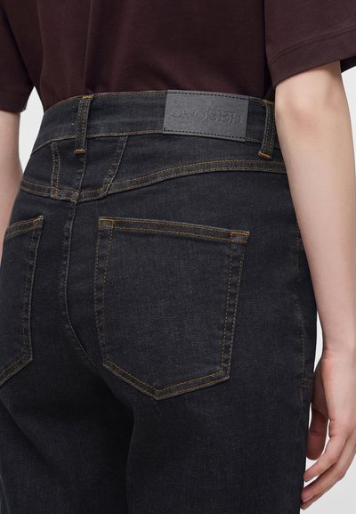 Μαύρο τζιν από denim με κίτρινη ραφή, που φοριέται από άτομο με σκούρο καφέ πουκάμισο, δείχνοντας τις πίσω τσέπες και μια ετικέτα με την επωνυμία που γράφει "CLOSED."