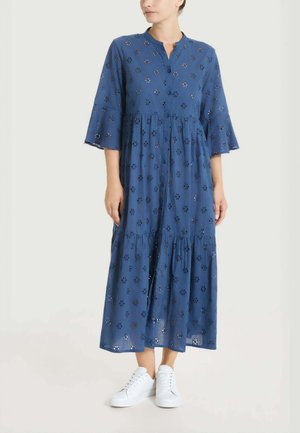 Donna che indossa un vestito midi blu con bottoni sul davanti, con ritagli floreali traforati e maniche a campana 3/4, abbinato a sneakers bianche.