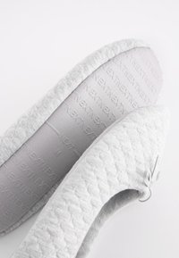 Hellgrauer Hausschuh mit strukturierter Oberseite, abgerundeter Zehenform und weichem Innenfutter. Verziert mit einer kleinen dekorativen Schleife an der Seite.