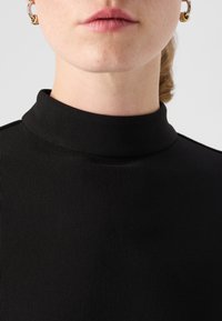 Čierny, rebrovaný rolák s priliehavým strihom. Detailný záber ukazuje hladkú textúru a vysoký golier. Viditeľné zlaté a strieborné kruhové náušnice.