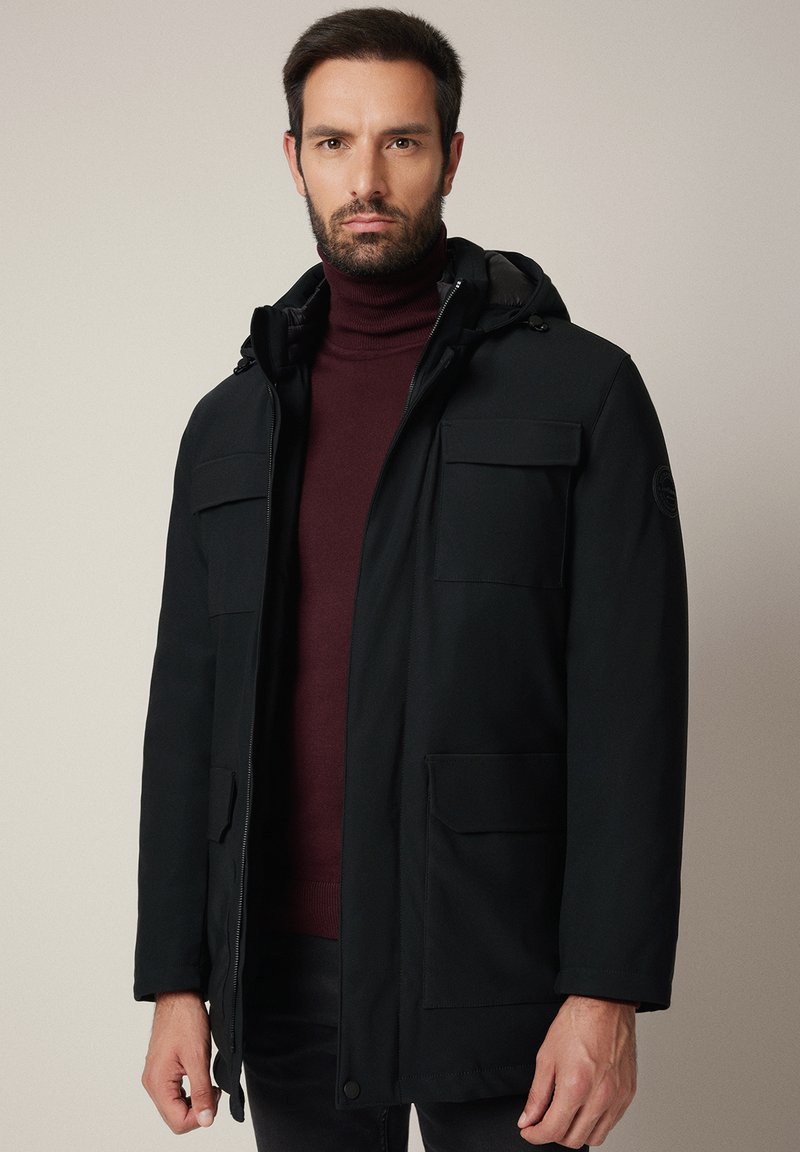 Czarna kurtka wierzchnia z kapturem, z zamkiem błyskawicznym z przodu, dwiema kieszeniami na piersi i lekko teksturowanym materiałem. Nosi się ją nad ciemnym golfem.