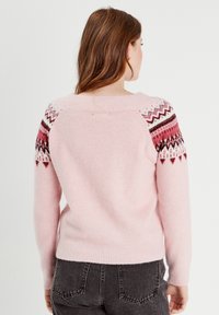 Jasnoróżowy sweter z wzorzystymi rękawami raglanowymi w kolorze czerwonym, białym i czarnym. Miękka tekstura, ściągacz przy kołnierzu i na hemie, luźny krój.