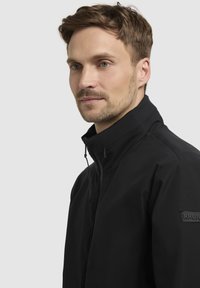 Chaqueta ligera negra con cuello alto, cierre de cremallera y cordón ajustable. Presenta un pequeño parche con el logotipo en la manga.