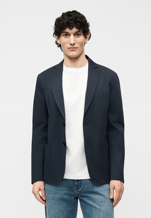 Jonge man met krullend haar, gekleed in een donker colbertje over een wit T-shirt en blauwe jeans, tegen een effen lichte achtergrond.