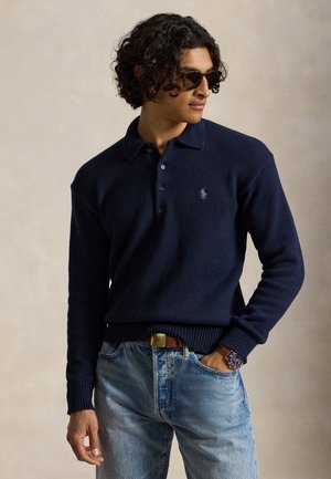 Homme aux cheveux bouclés portant un pull polo en tricot bleu foncé, un jean bleu clair, des lunettes de soleil et une montre en cuir marron, main dans la poche, regardant sur le côté.
