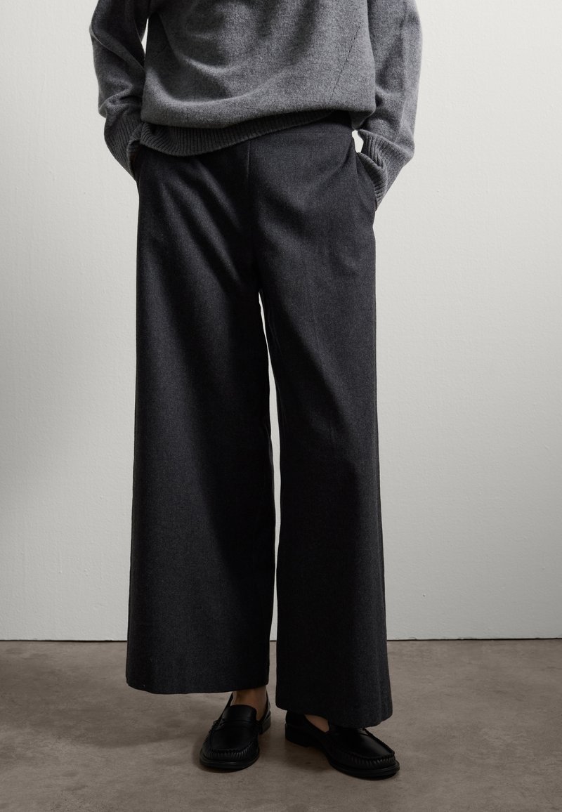 Pantaloni a gamba larga, di colore grigio scuro, realizzati in tessuto morbido, con una vestibilità comoda e tasche frontali, abbinati a mocassini neri.